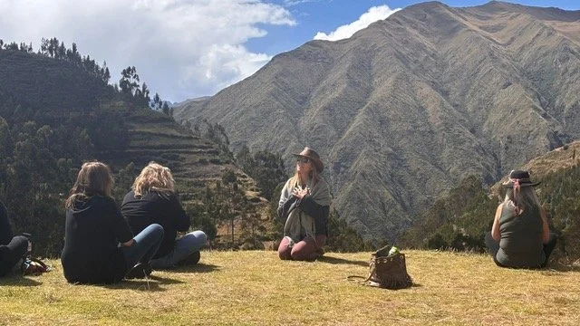 Four women sitting on grass in front of mountains, two facing away and two facing sideways, one woman appears to be speaking or leading an activity.