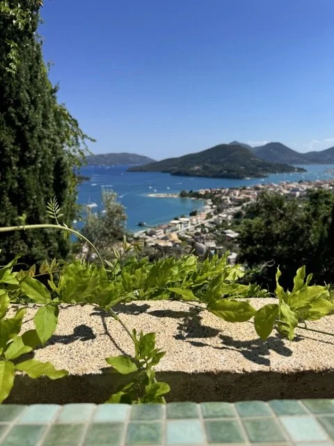 Scenic view of a coastal city with a lake, surrounded by green hills and mountains, under a bright blue sky.