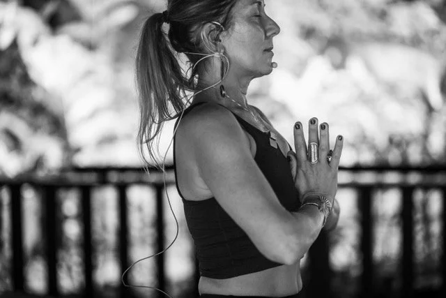 A woman practicing yoga outdoors, standing with her hands pressed together in prayer position, wearing a tank top and jewelry.