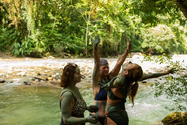 Three women standing in a river, celebrating and raising their arms, surrounded by lush green trees.