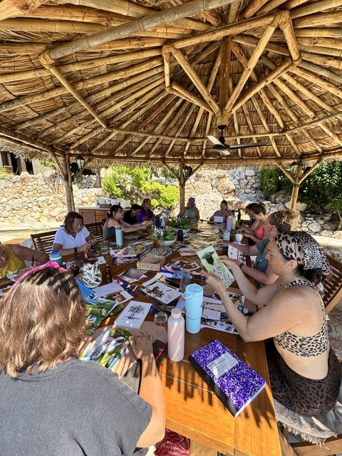 Group of women sitting around a long table under a thatched roof, engaging in a crafting or discussion session, with papers, books, and drinks on the table.