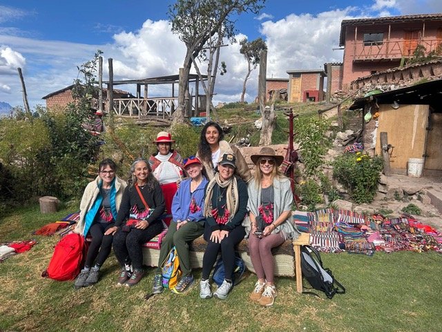 Group of women sitting and standing outdoors in a rural setting, with colorful woven mats and bags, trees, and rustic buildings in the background.