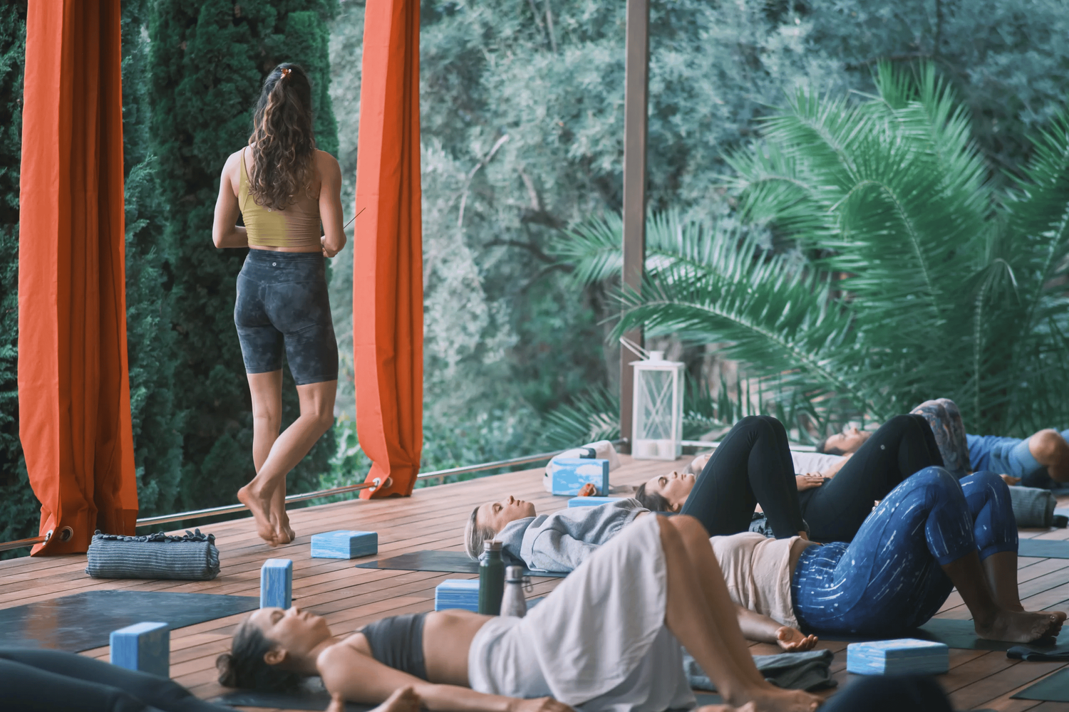 A yoga class taking place in a room with large windows overlooking a lush green forest. Several women are lying on mats doing relaxation poses, while a woman at the front appears to be instructing or observing.