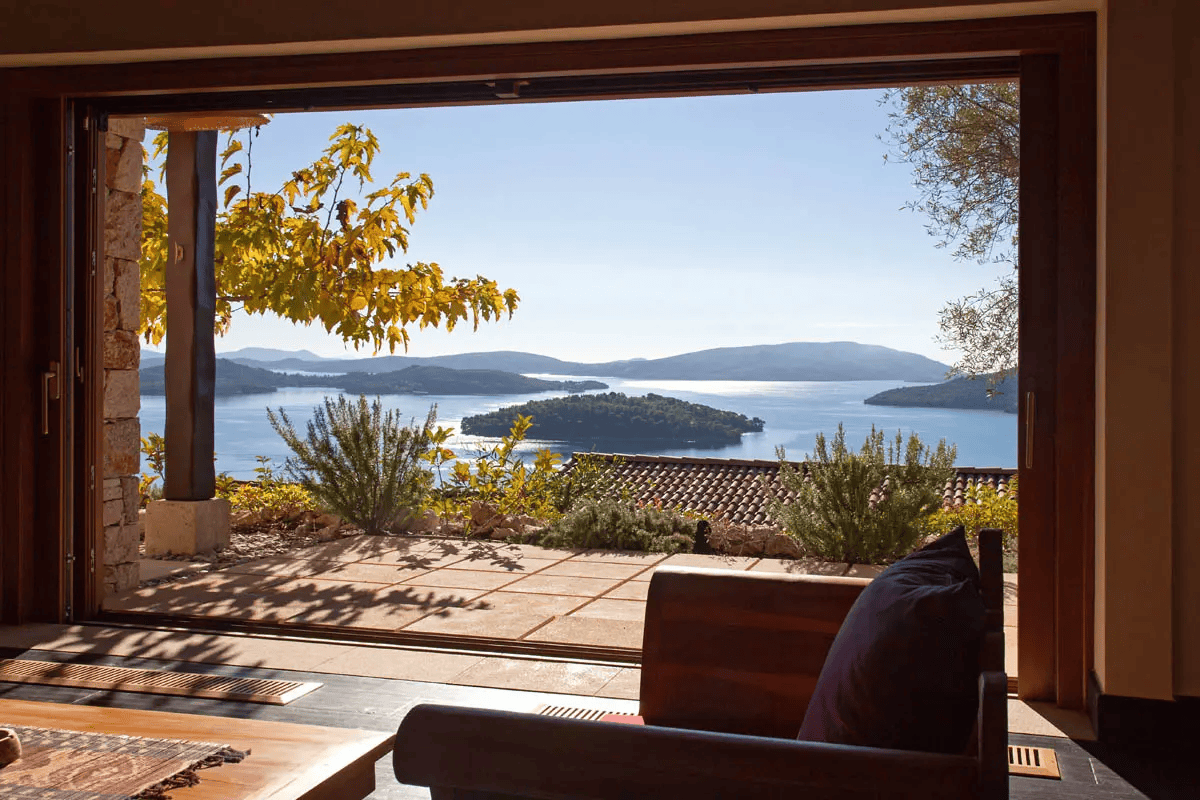 View of a lake and distant hills seen through an open sliding glass door from inside a house, with outdoor plants and roof visible.
