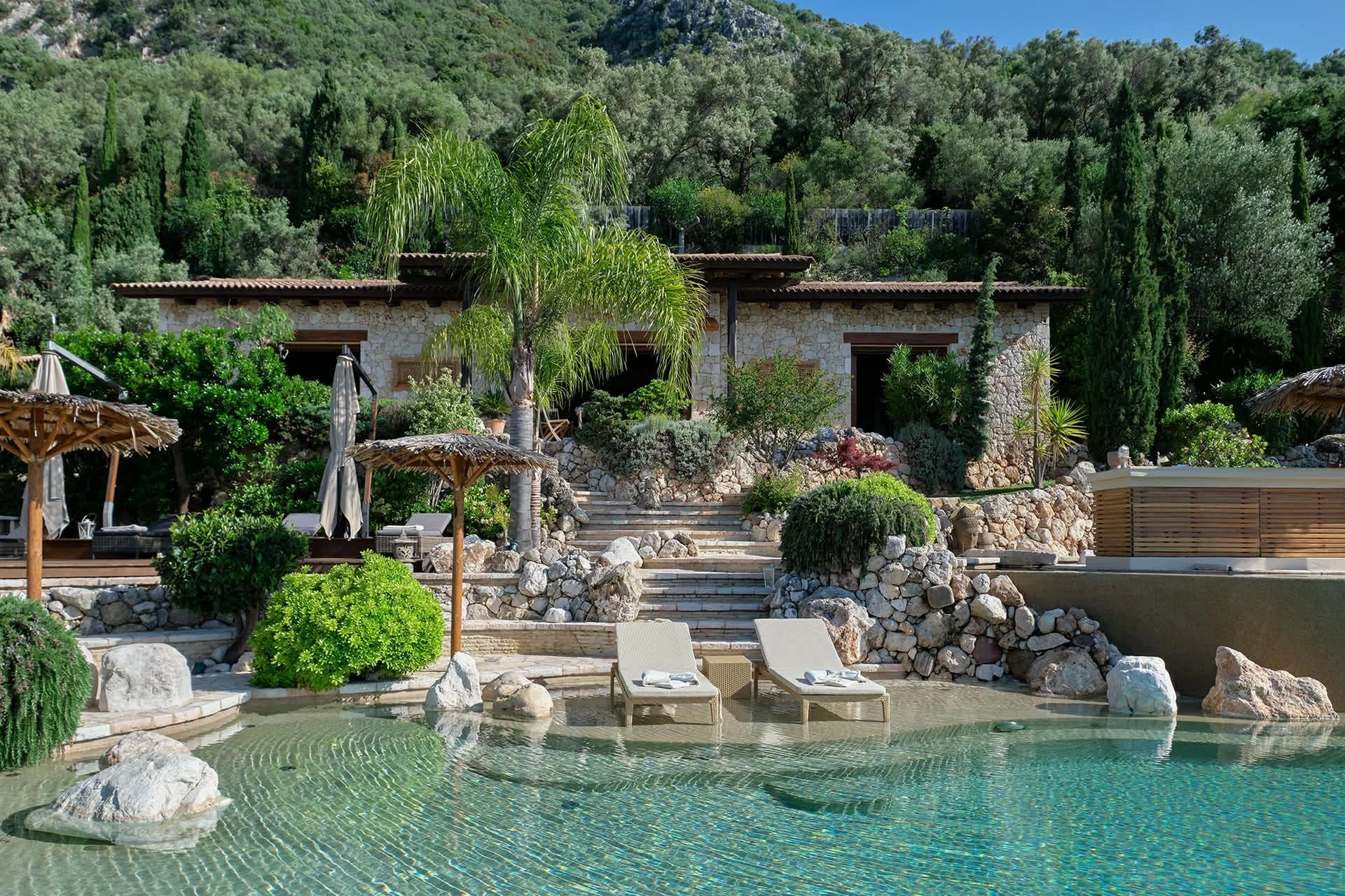 A luxurious outdoor swimming pool with clear water, surrounded by rocks, trees, and lush green plants. There are two lounge chairs with towels and a couple of shaded cabanas nearby, with a stone house and a hillside covered in trees in the background.