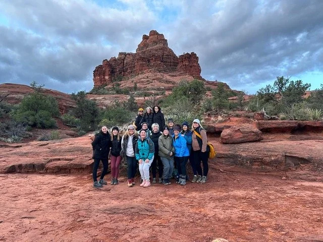 Group of people hiking in a desert landscape with red rock formations and cloudy sky.