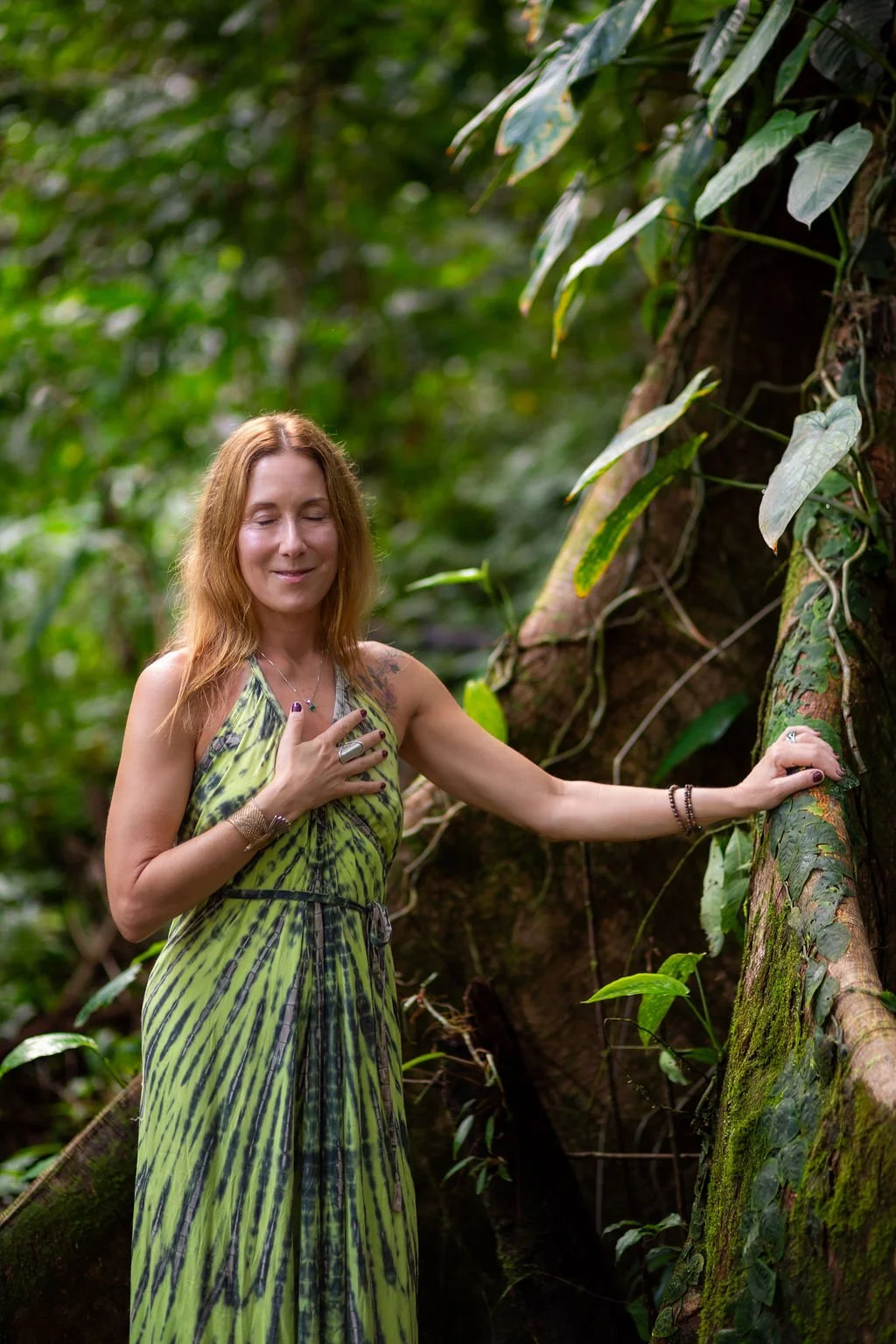 A woman with long reddish hair and a peaceful expression stands in a lush green jungle, wearing a green and black patterned dress, with her hand on her chest and the other on a moss-covered tree.