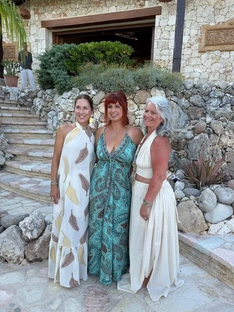Three women standing outdoors in front of rocks and plants, smiling and posing for a photo, dressed in summer dresses.