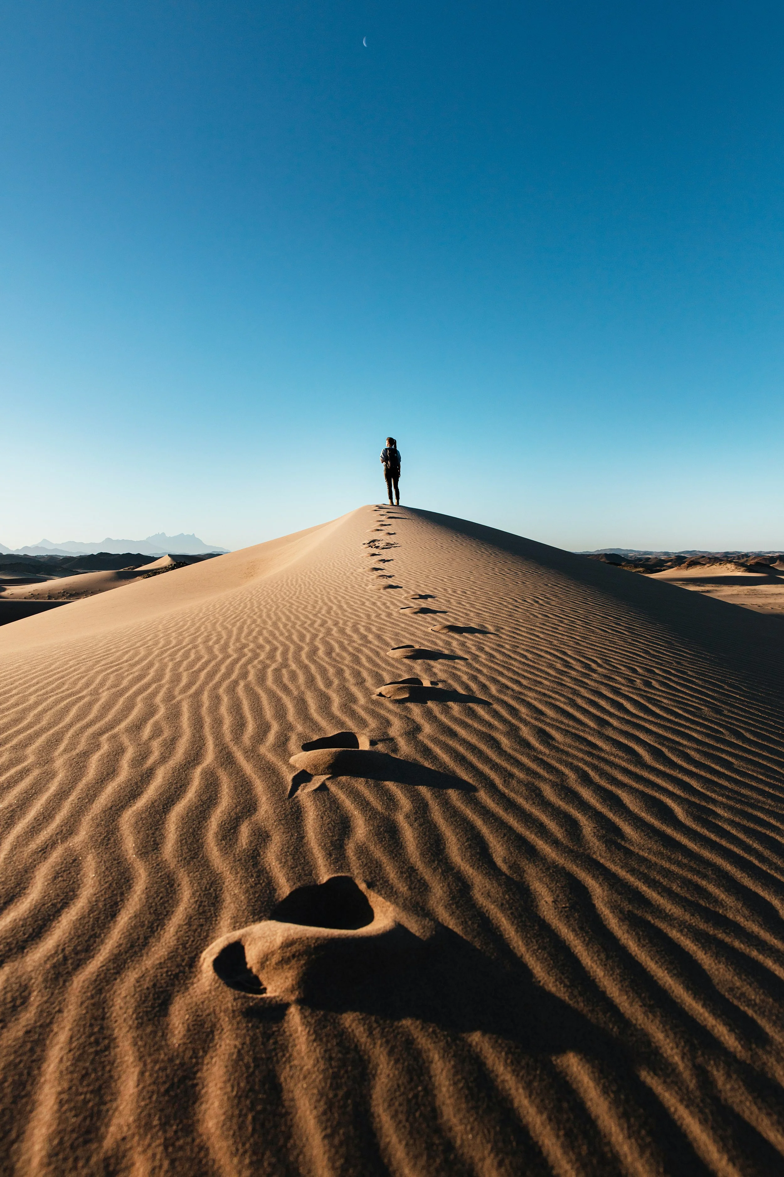 A person standing alone on top of a sand dune in the desert, with footprints leading up to them and a crescent moon in the clear blue sky.