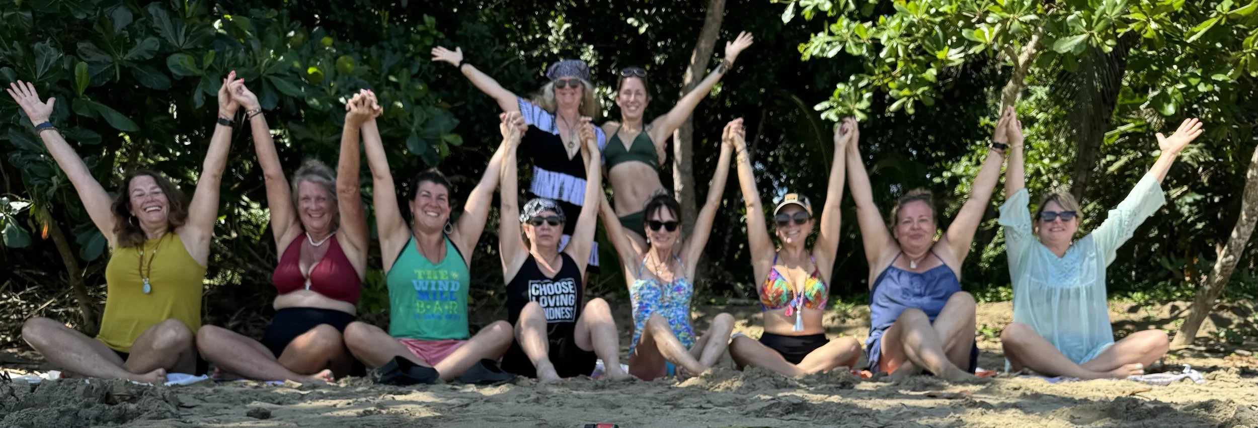 Group of women sitting on the beach with their legs crossed and arms raised, smiling and enjoying a sunny day surrounded by green trees.