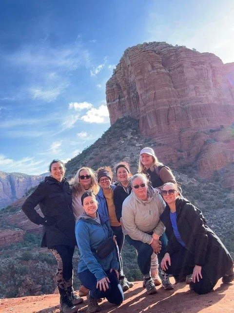 Group of eight women posing outdoors in front of a large red rock formation against a blue sky.