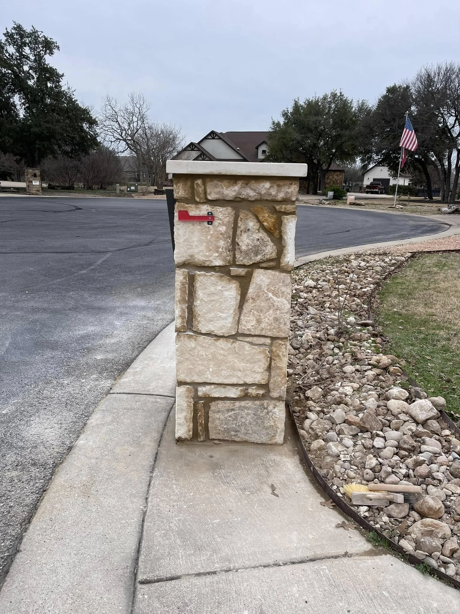 A stone mailbox post with a red flag on the left side, situated on a sidewalk next to a curved road, with a yard containing gravel and rocks on the right and residential houses in the background, and an American flag on a pole to the right.