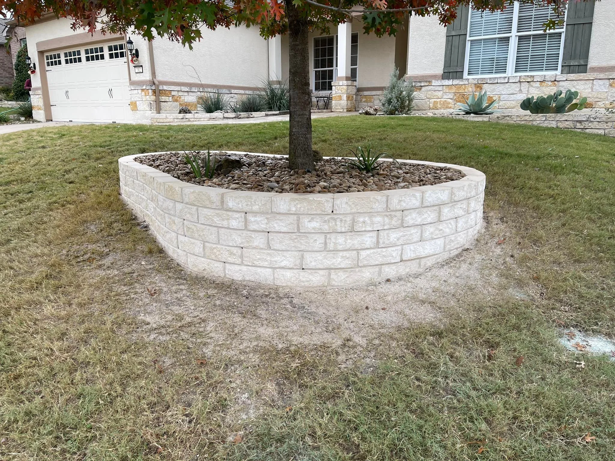 A landscaped front yard with a tree in a circular brick planter filled with rocks, in front of a house with a garage, stone and stucco exterior, and various plants.