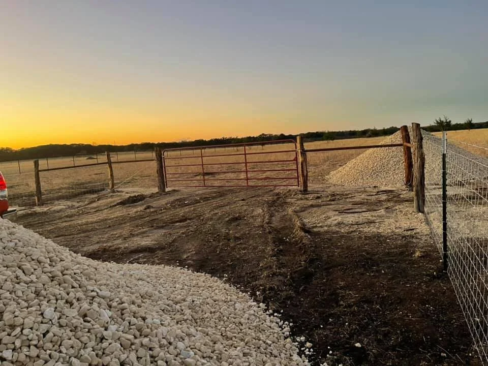 A rural farm entrance with a closed red metal gate, dirt driveway, gravel piles on both sides, and fencing along the property boundary during sunset.