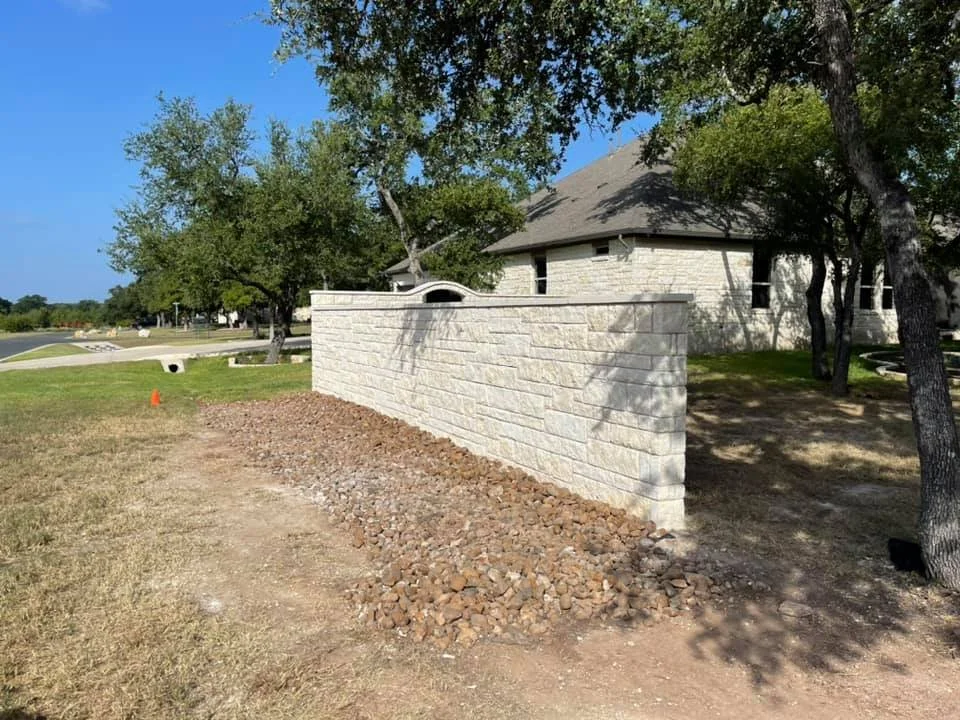A stone wall in front of a house with trees and a grassy area around, under a clear sky, with some gravel and dirt in the foreground.