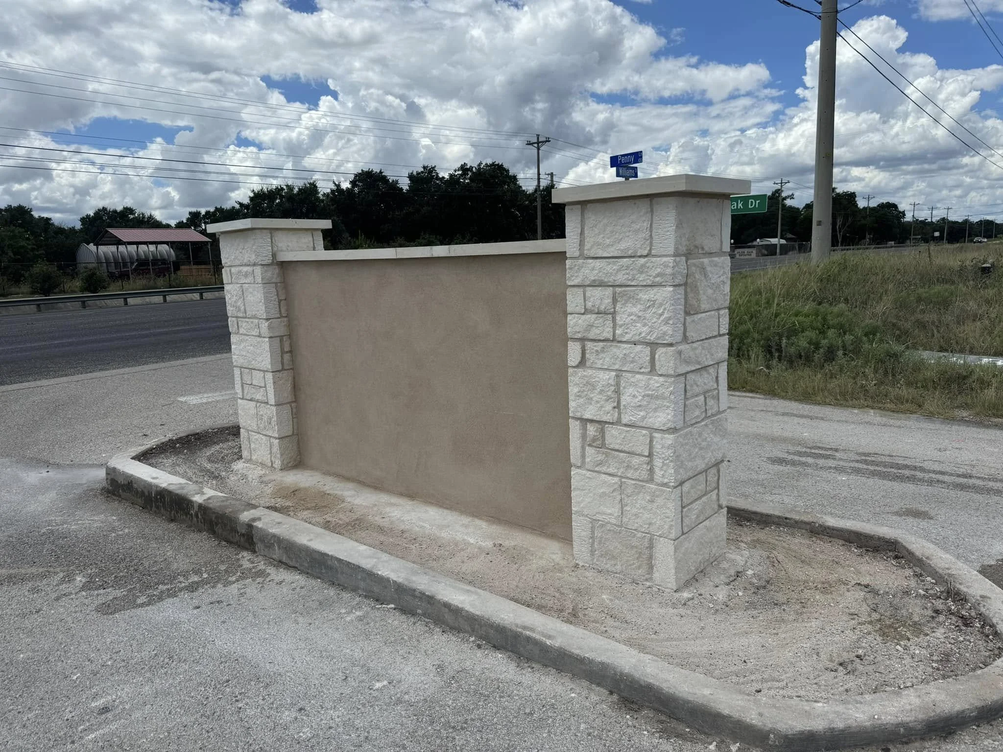 A newly constructed stone and stucco fence or wall on a roadside, with street signs and power lines in the background. The sky is partly cloudy.