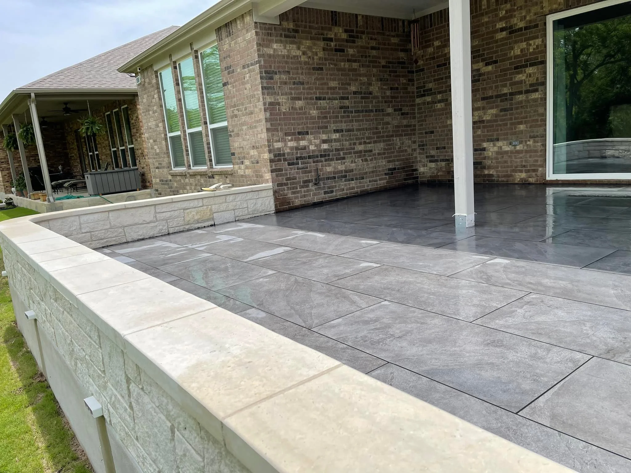 View of a house's back patio with tiled flooring, brick walls, and sliding glass door. The patio has a white stone curb and is surrounded by a grassy yard.