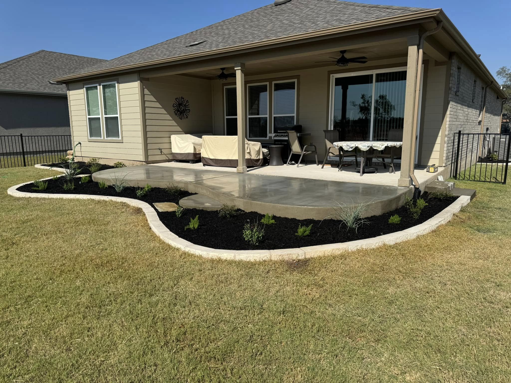 Backyard patio with a concrete deck, surrounded by a curved garden bed with mulch and small plants, and a well-maintained grassy lawn.
