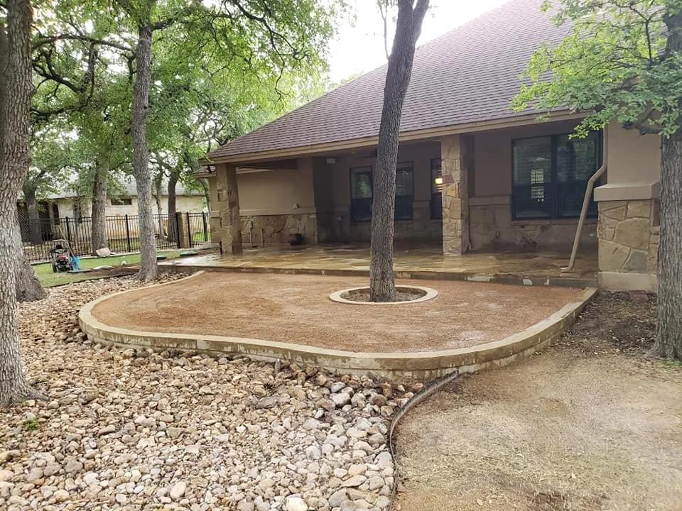 Backyard with a newly landscaped area featuring a brick border, a central tree, and a patio under a house's covered porch surrounded by trees and rocks.