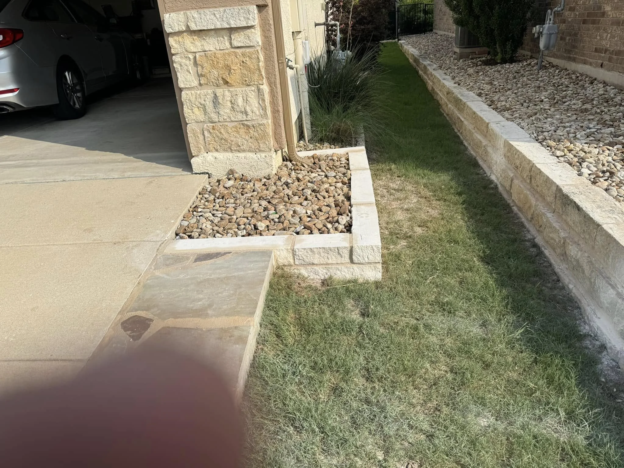 A driveway leading to a garage with a parked silver car, bordered by a stone and gravel landscape with a small grassy area, a brick wall, and some plants.