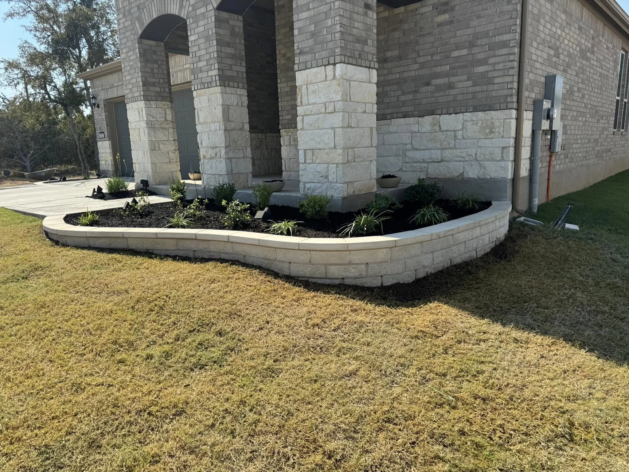 A landscaped area around the corner of a brick house with a curved stone flower bed filled with plants and mulch, situated on a lawn.