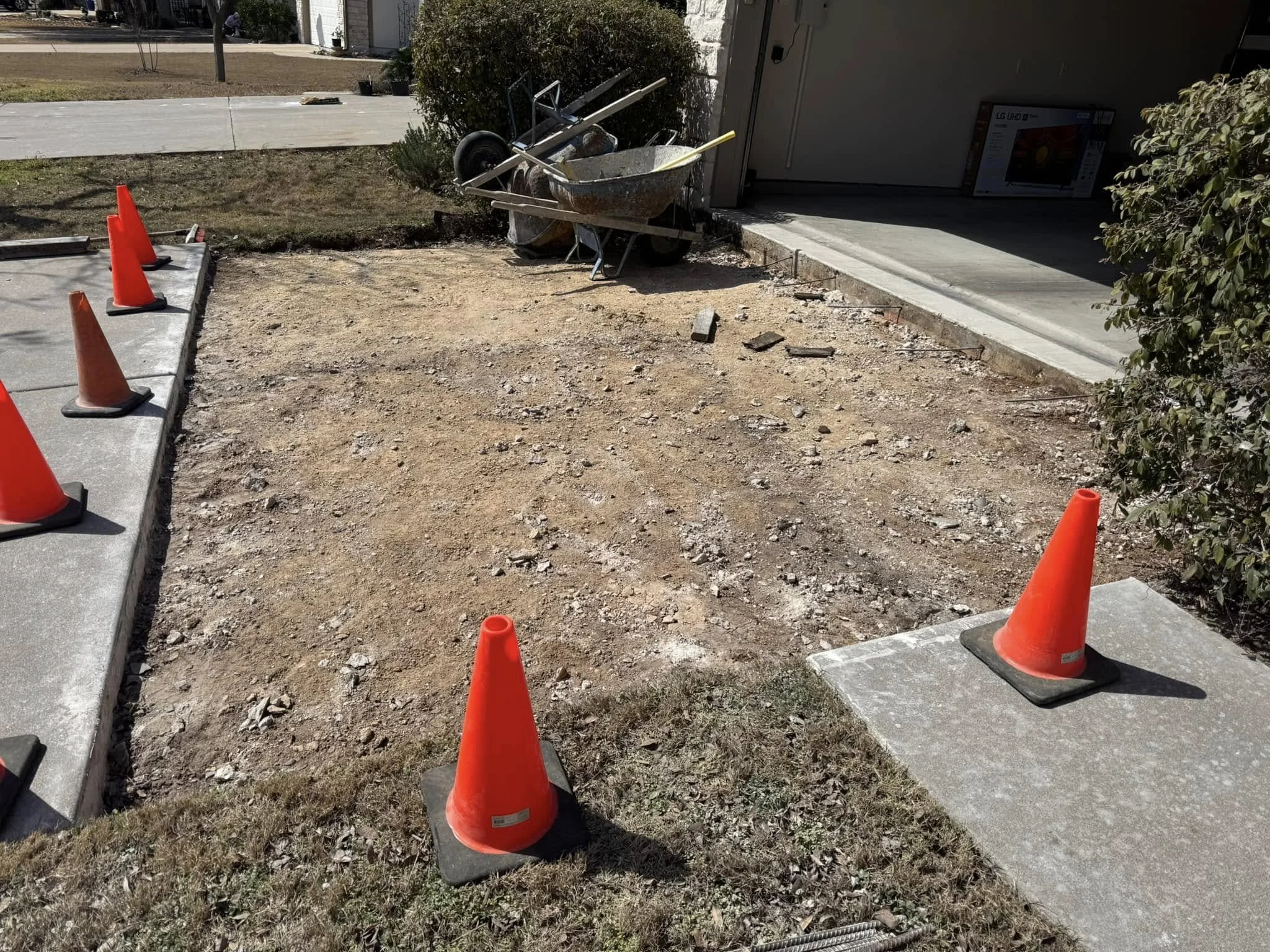 Driveway under construction with orange traffic cones, concrete sidewalk, a wheelbarrow, and construction tools near the house.
