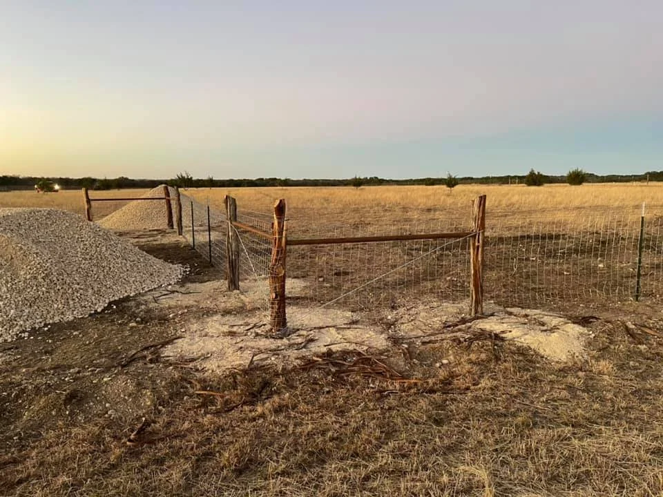 A rural landscape with a partially built fence, a pile of gravel, and a wide open field with a few trees in the background at sunset.