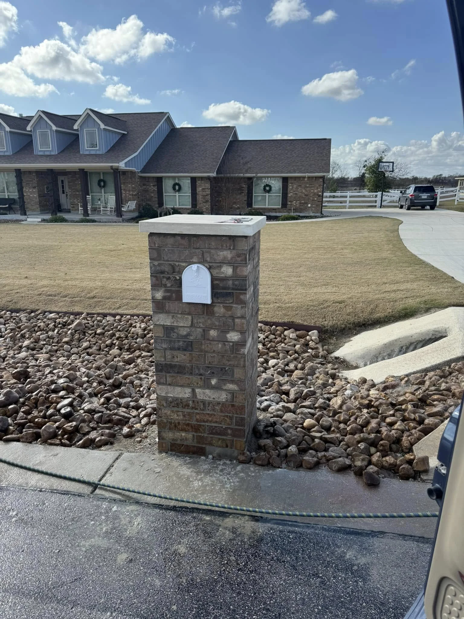 View of a brick mailbox in the front yard of a house with a lawn and driveway, under a partly cloudy sky.