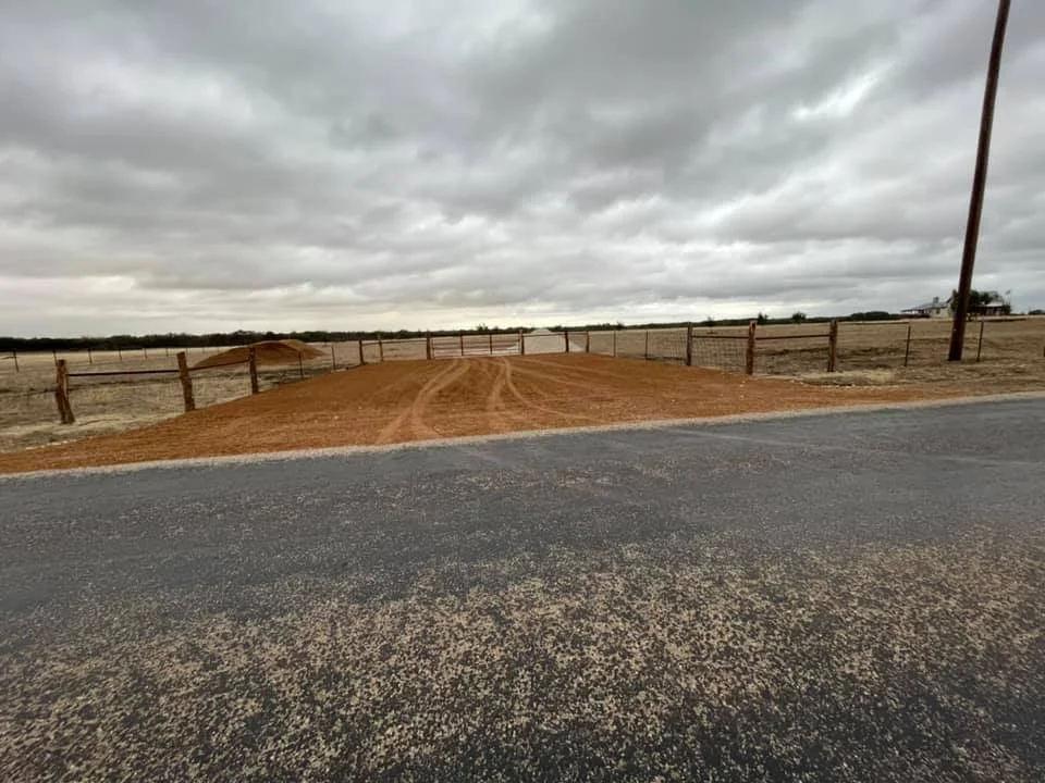 A rural landscape with a dirt driveway leading to open land, a wooden fence, cloudy sky, and a paved road in the foreground.
