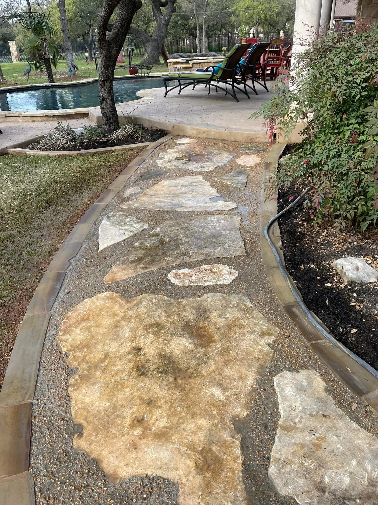A stone walkway leading from a backyard patio to a swimming pool area, bordered by a curved wooden edge and surrounded by grass and shrubs, with patio chairs and a table in the background near a pool.