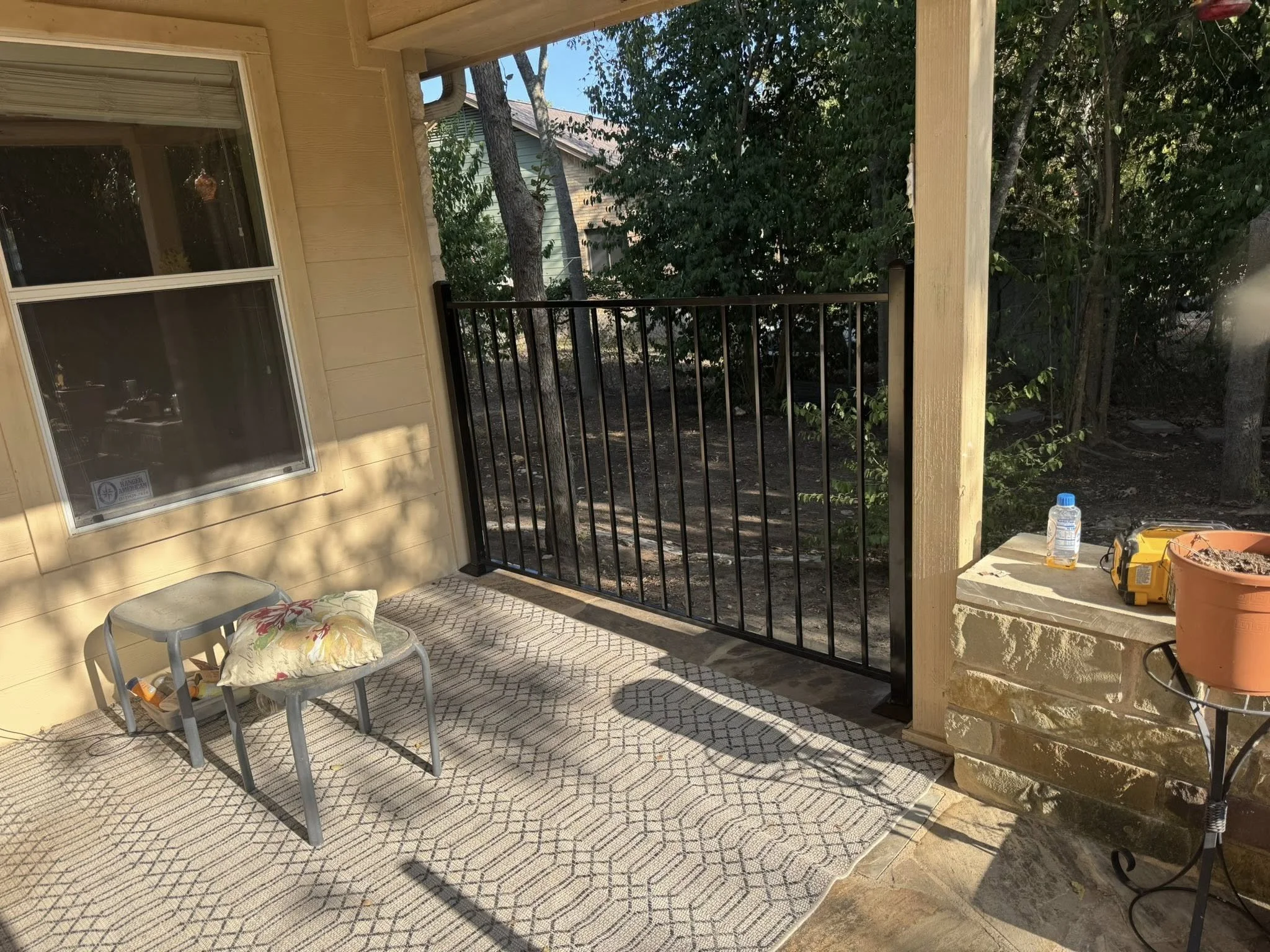 A porch with a beige wall, a window, a small table with a pillow, and a railing. There are trees and foliage outside, with sunlight casting shadows on the ground and wall. A grow light, a bottle, a tool, and a plant pot are on the right side.