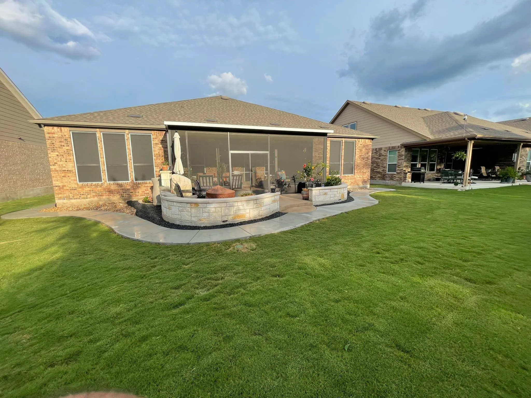 Backyard with a brick house featuring a screened-in porch, paved walkway, green lawn, and neighboring houses in the background under a partly cloudy sky.