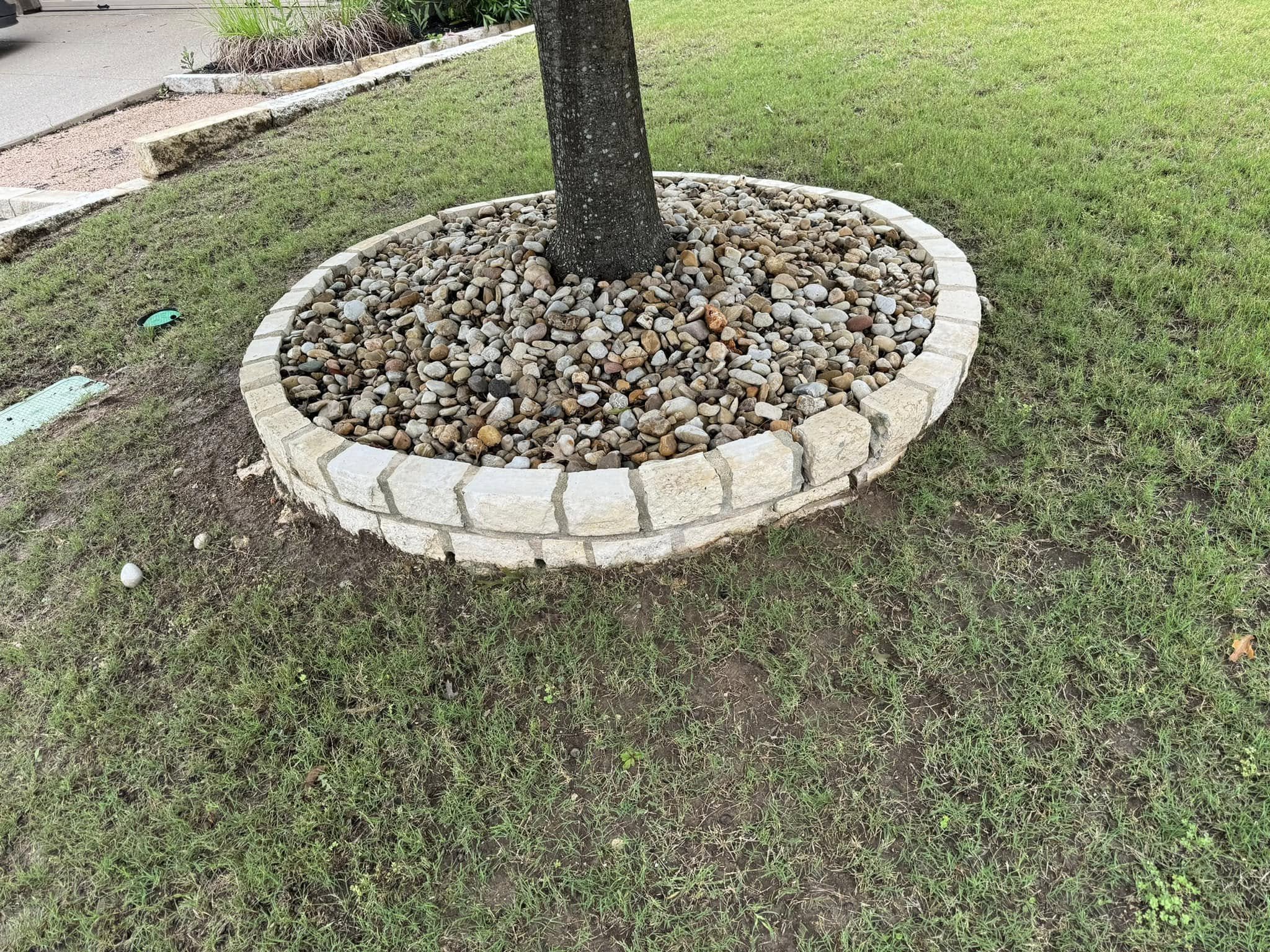 A round garden bed with a tree in the center, surrounded by a border of light-colored bricks. The bed is filled with small, rounded stones. The surrounding grass is green, and part of a sidewalk and a small landscaped area are visible in the backgrou