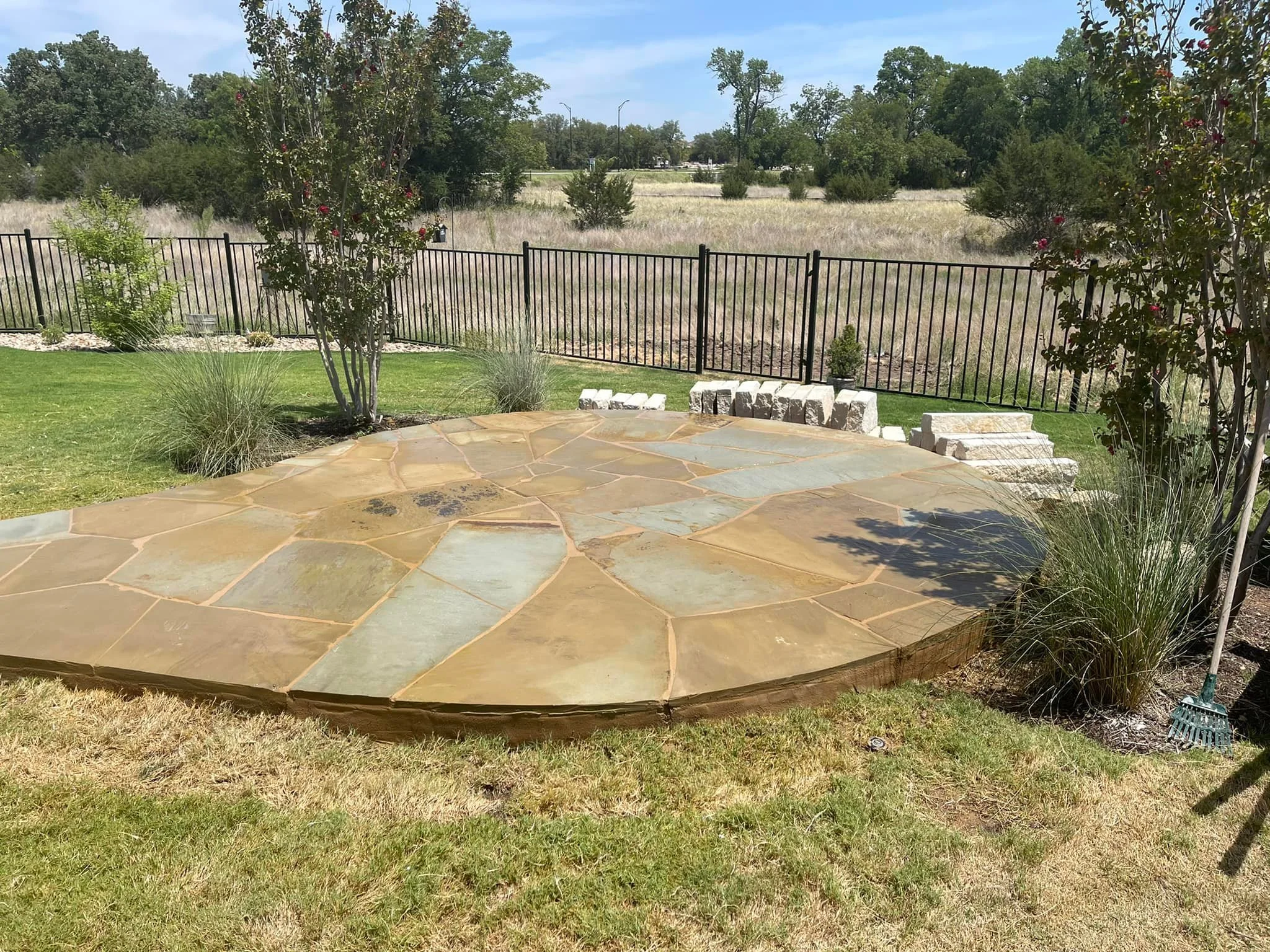 Backyard patio with large irregular flagstone surface, surrounded by grass, trees, and bushes, fenced with a black metal fence, and a partly cloudy sky.