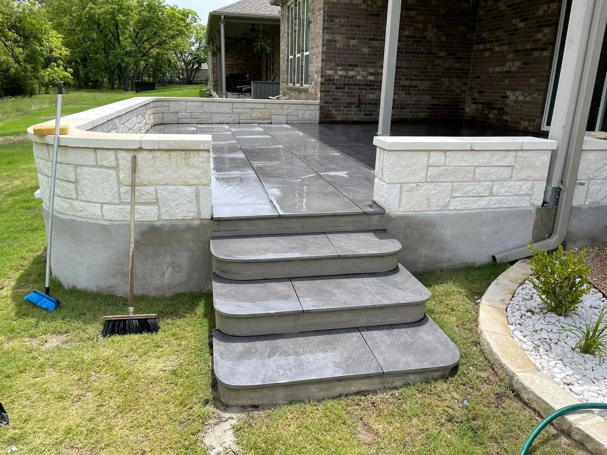 A newly constructed outdoor patio with gray tile flooring and a short staircase leading up to a brick house. There are tools like brooms nearby and a decorative flower bed with white rocks to the right.