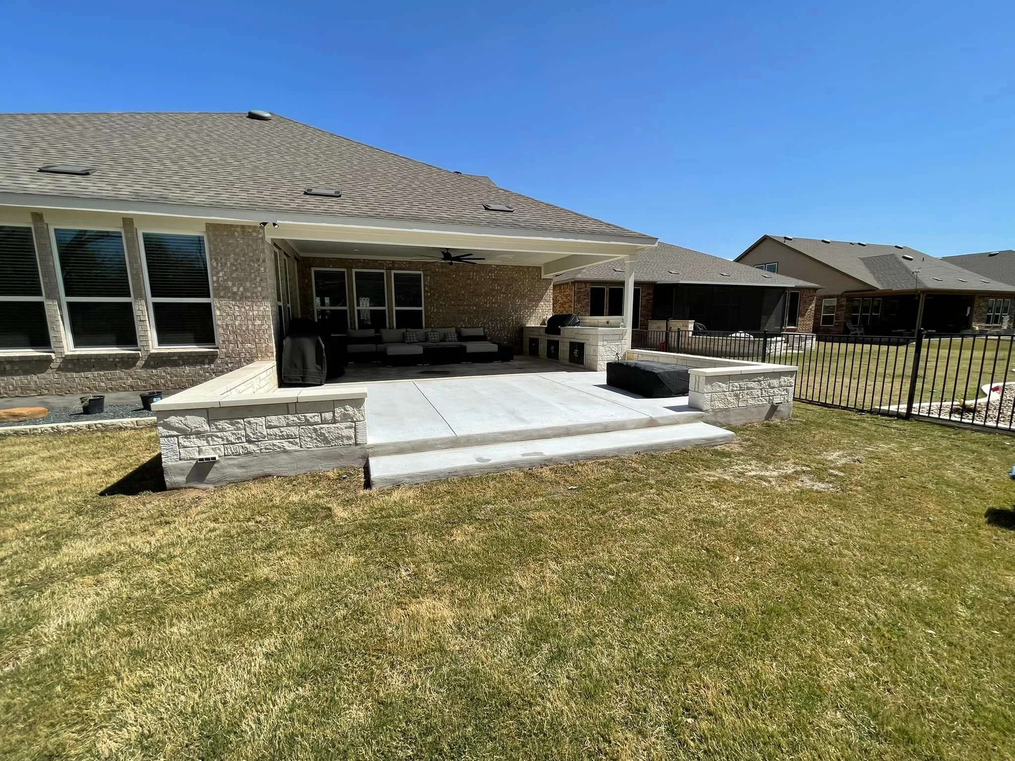 Backyard patio with a raised concrete platform, outdoor seating, grill, and partial brick wall, enclosed by a black metal fence, with neighboring houses in the background, under a clear blue sky.