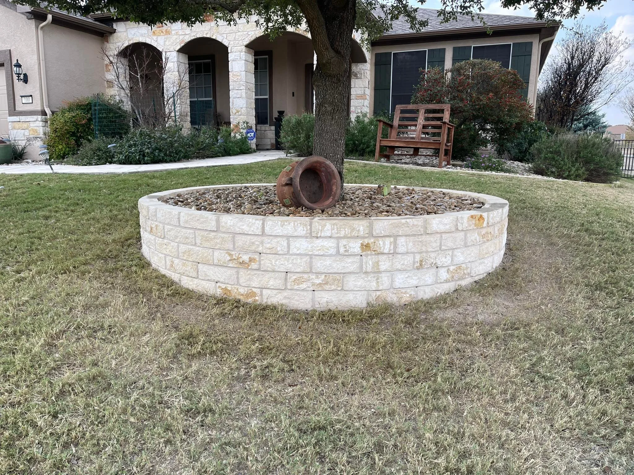 Front yard of a house with a circular brick planter containing a large tree, rocks, and an overturned clay pot. There is a wooden bench and various bushes and plants in the background.