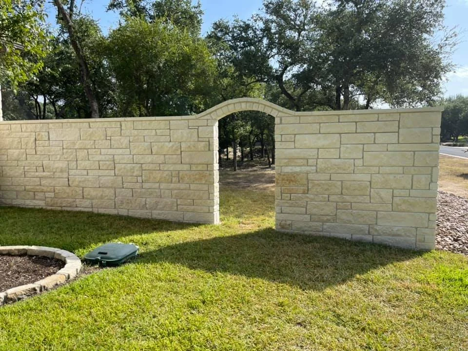 A beige brick wall with an archway in the middle, located in a grassy yard with trees in the background and a sidewalk to the right.