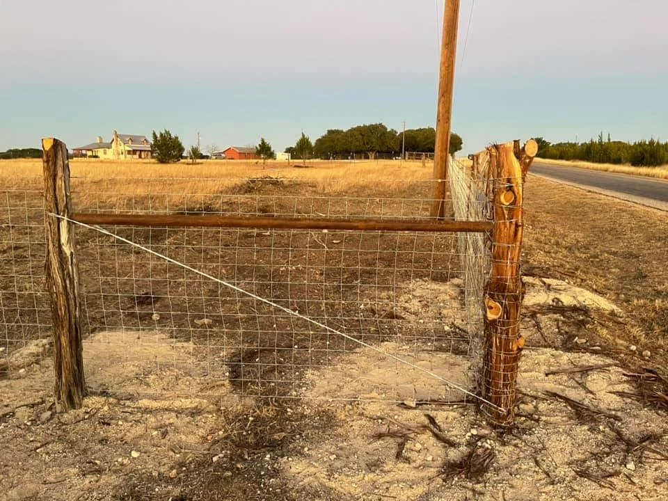 A rural fenced area with a wooden gate, dirt ground, and a country road to the right. In the background, there are houses and trees under a clear sky.