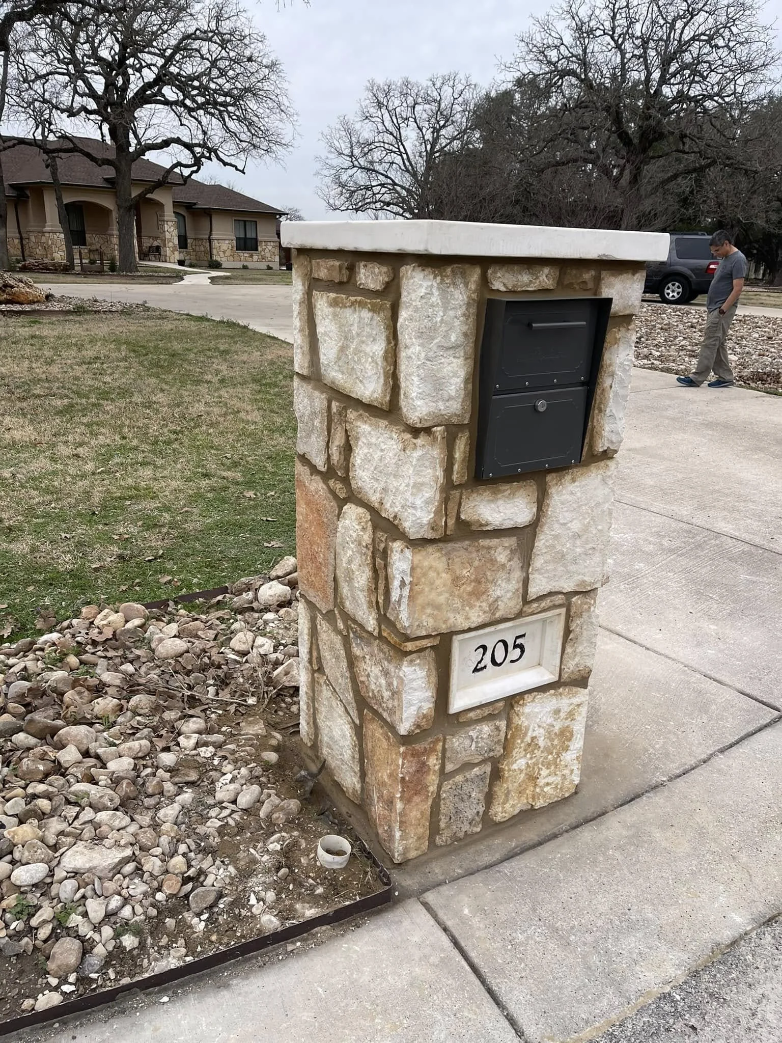 Stone mailbox with house number 205 on a sidewalk in a suburban neighborhood with trees and houses in the background.