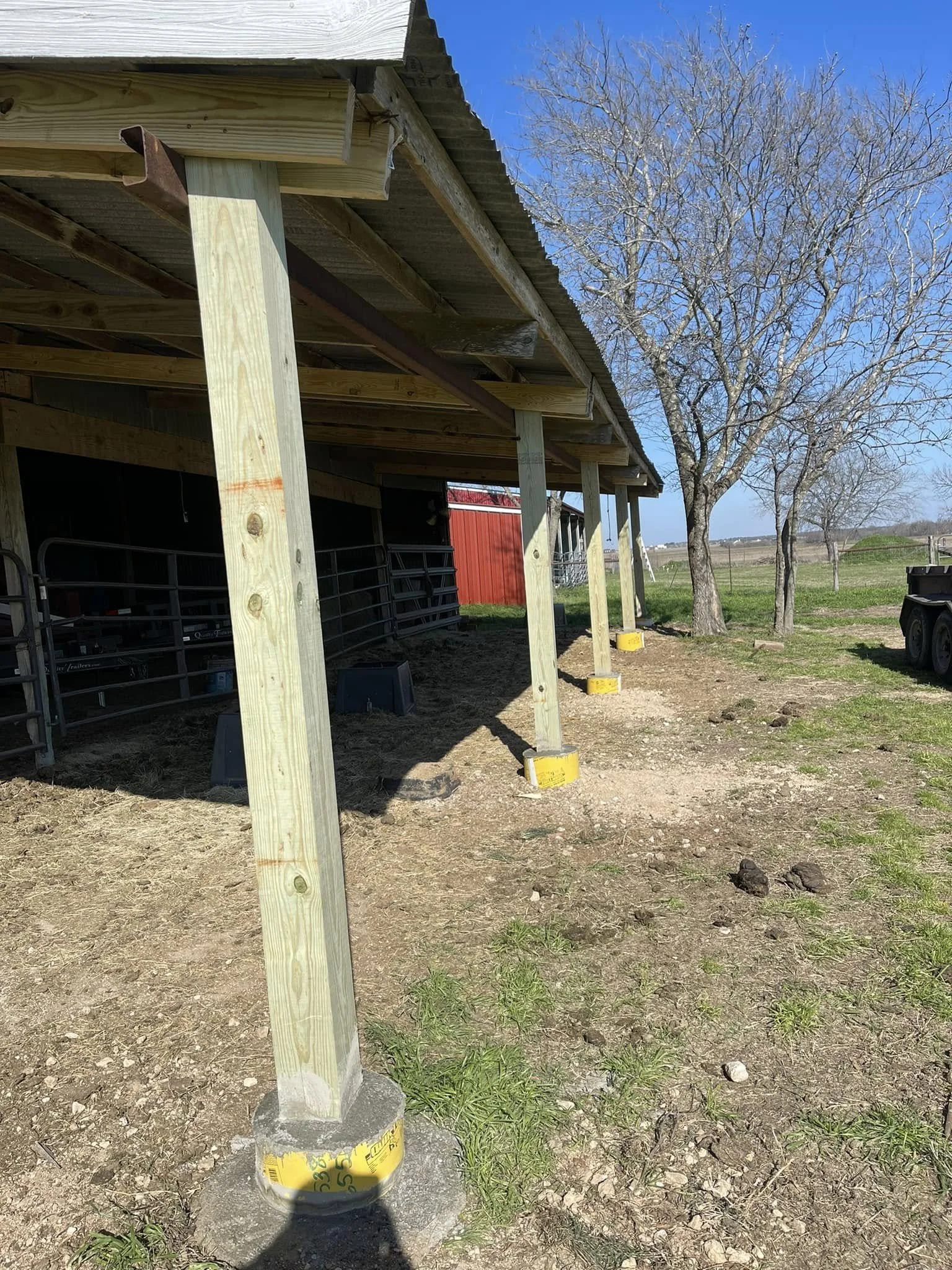 Construction of a wooden porch or deck supported by vertical wooden posts with concrete foundations, located on a farm or rural area with trees and open fields in the background on a sunny day.