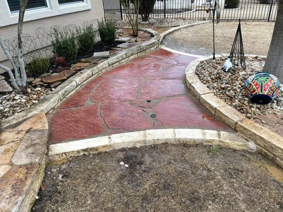 A winding red flagstone pathway in a backyard, bordered by a stone curbing, with a gardening bed on one side containing plants, rocks, and decorative items including a colorful spherical ornament.