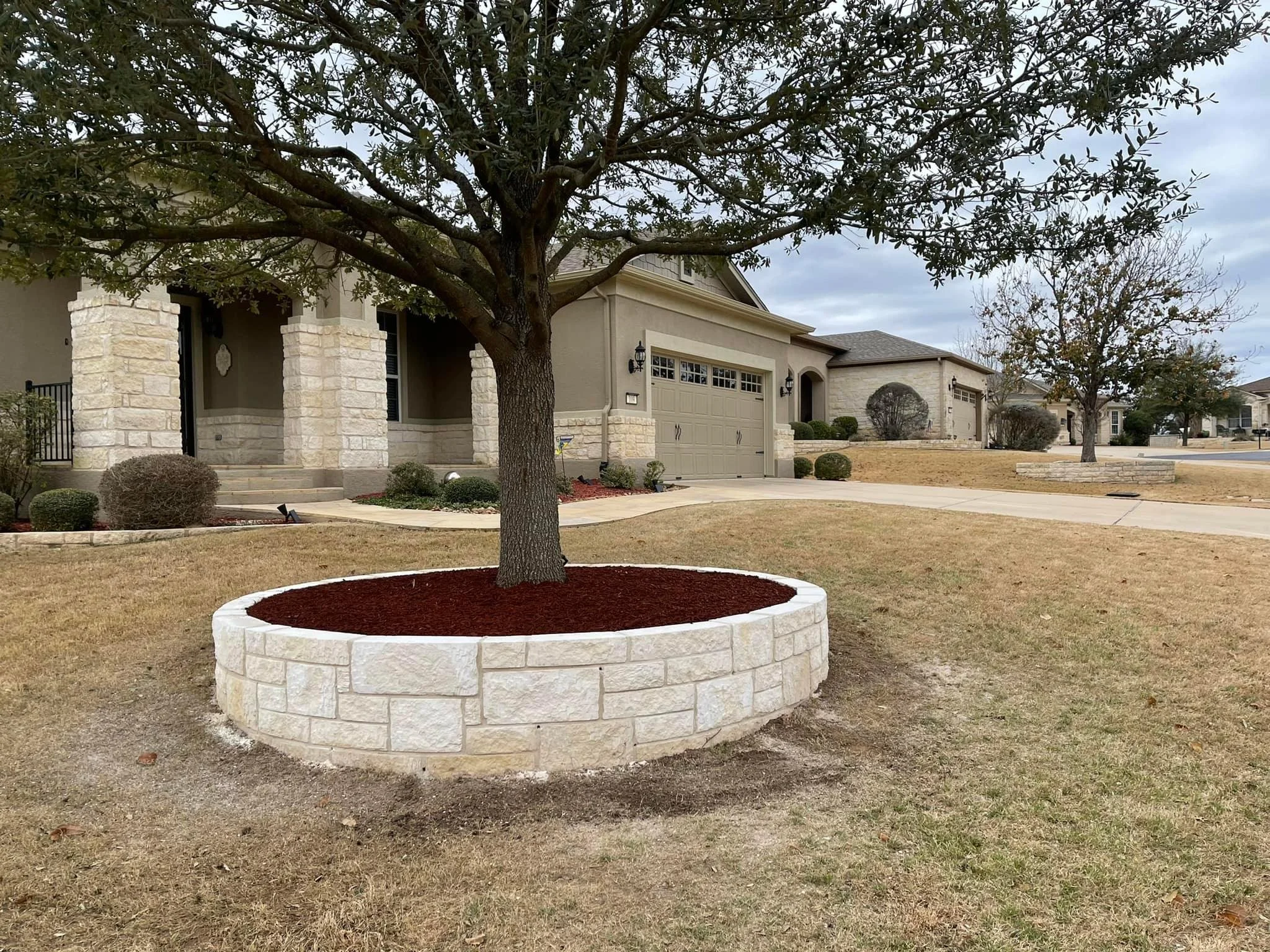 A suburban house with a front yard featuring a tree with a circular stone planter, a concrete driveway, and multiple trees and bushes in the yard under an overcast sky.