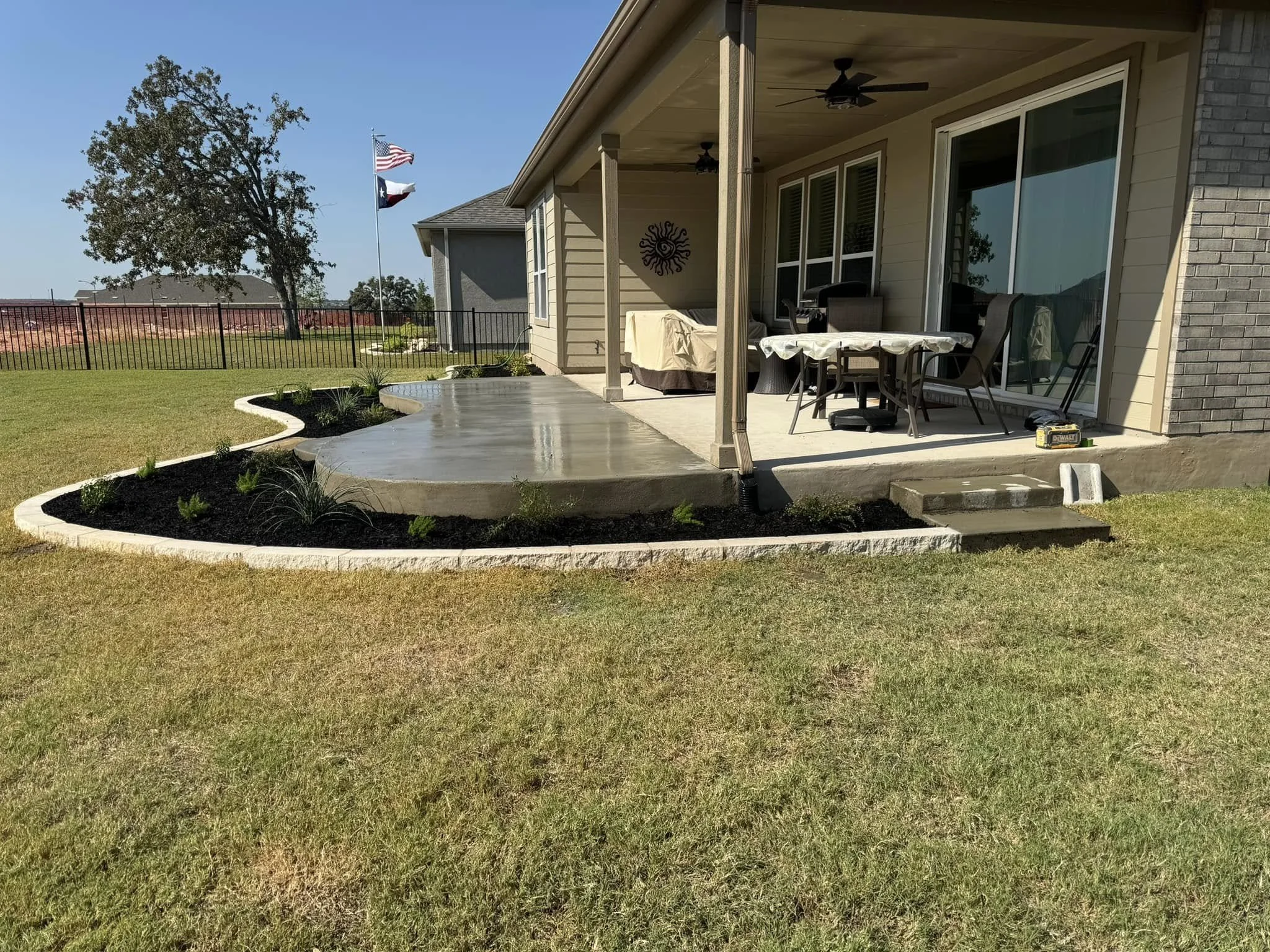 Backyard patio with freshly poured concrete, outdoor furniture, and a fenced yard with trees and an American flag
