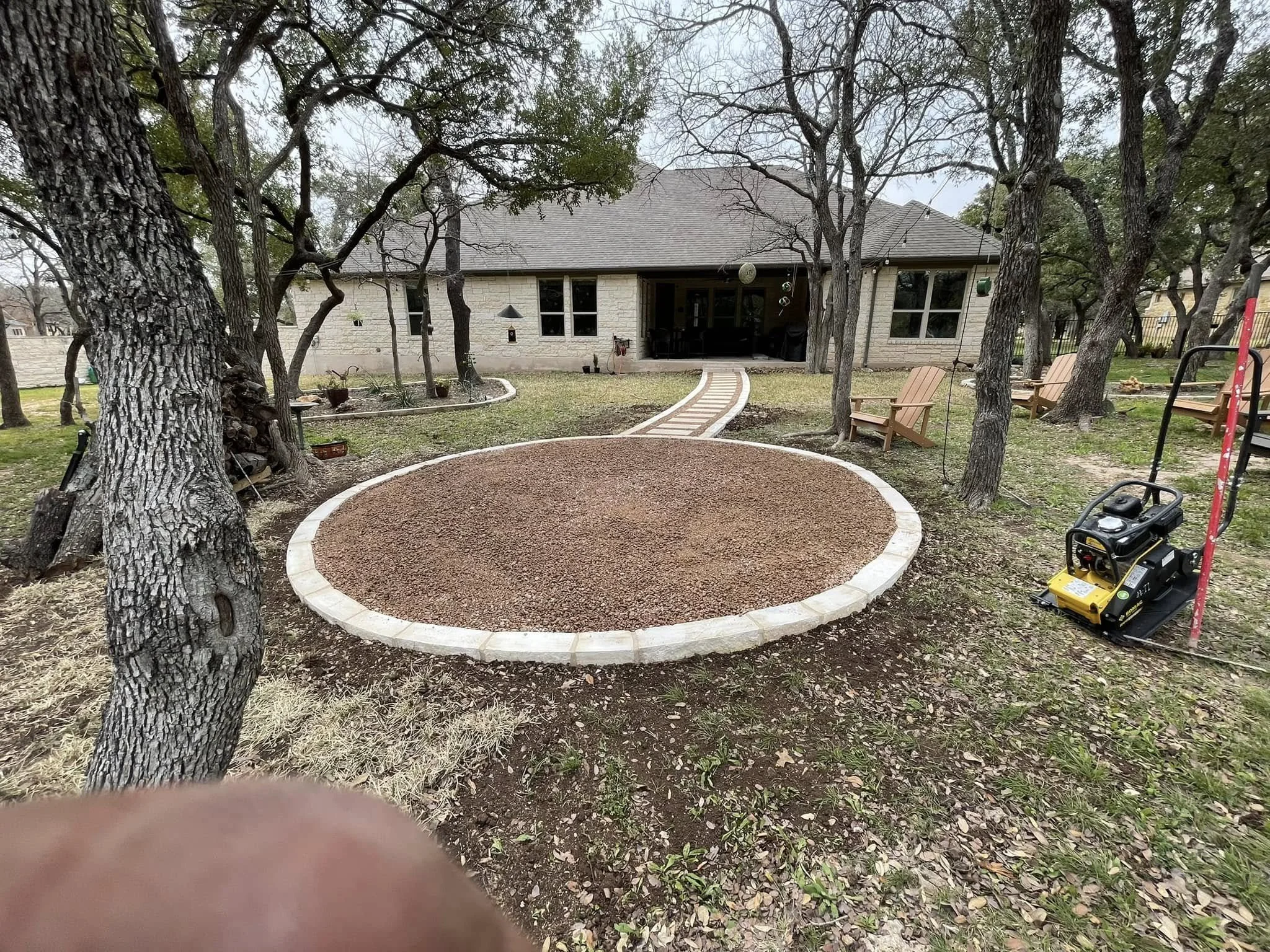 Backyard with a newly constructed circular gravel patio surrounded by a white brick border, a brick pathway leading to the house, several trees, wooden chairs, and gardening equipment.