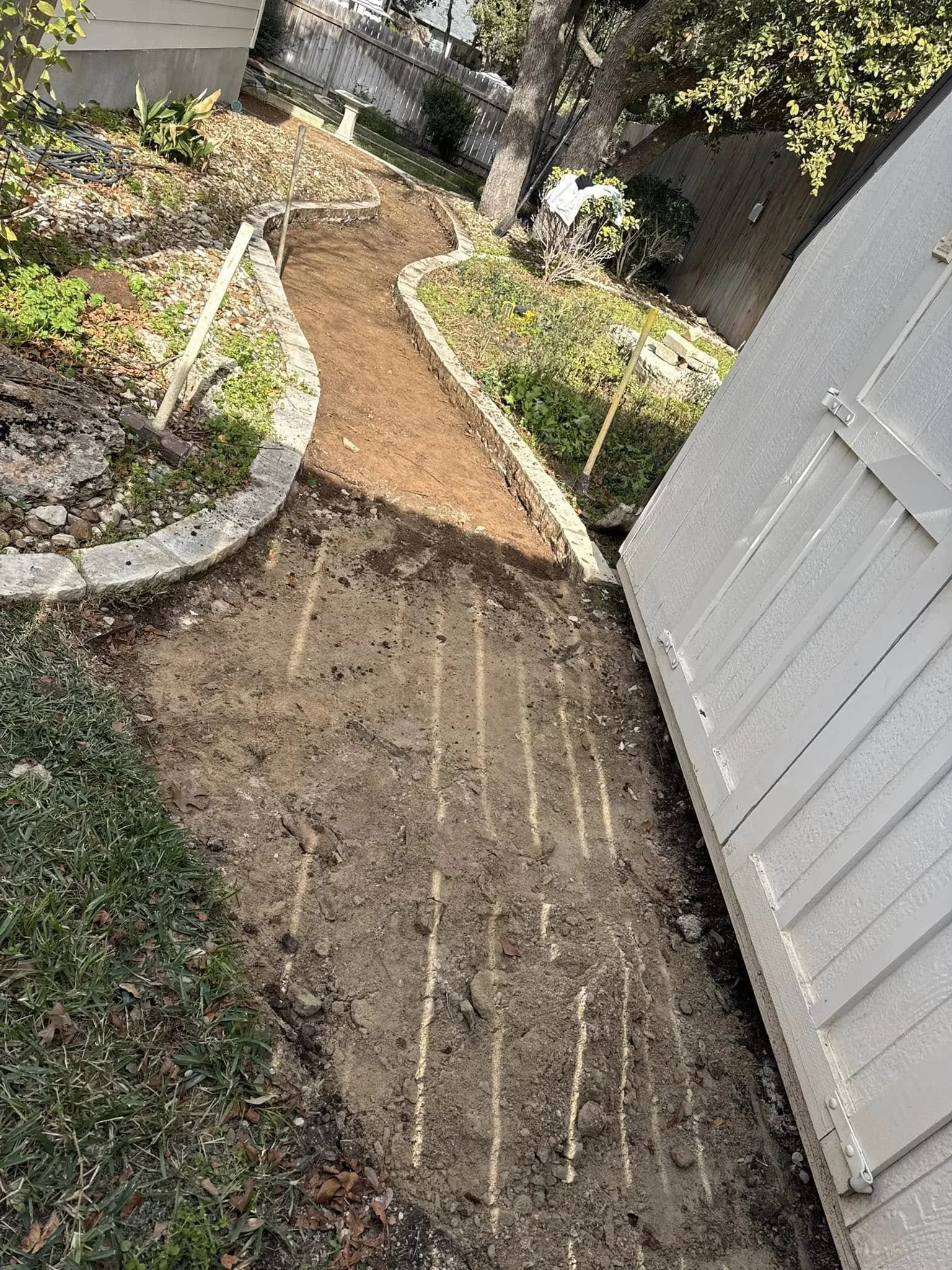 A backyard garden with a pathway under construction. The pathway is partially dug out with visible lines for future pavers. There are garden beds with plants and flowers on either side, and a white shed or building on the right. A wooden fence and tr