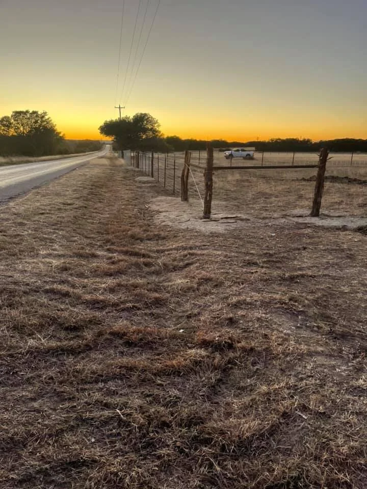 A rural landscape at sunset with a dirt road on the left, a wire fence on the right, a few trees, and a white truck in the distance.