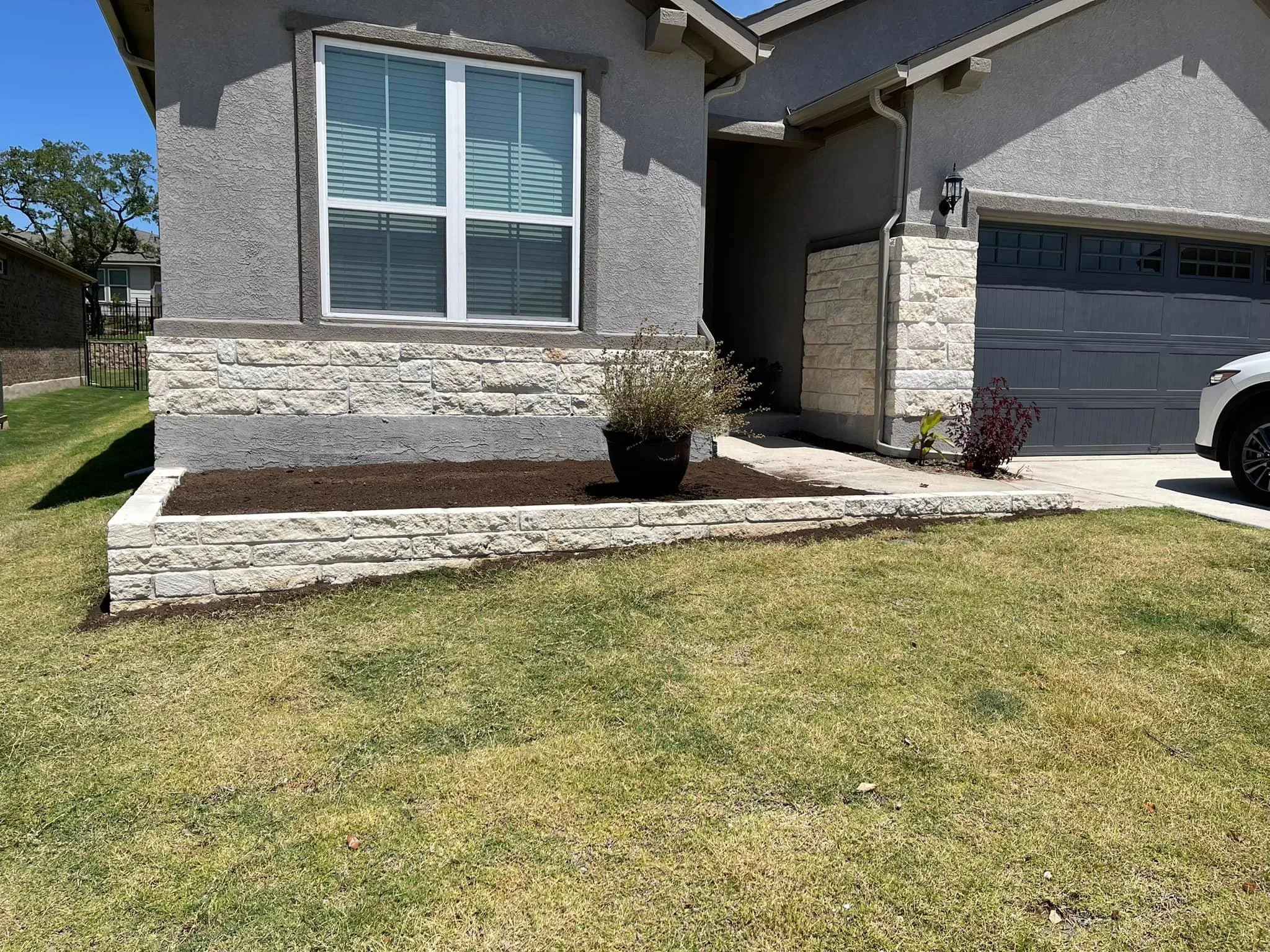 Front side view of a house showing a flower bed with fresh soil, a potted plant, and a large window with closed shutters. The house has gray walls with stone accents, a gray garage door, and a white car parked in the driveway.