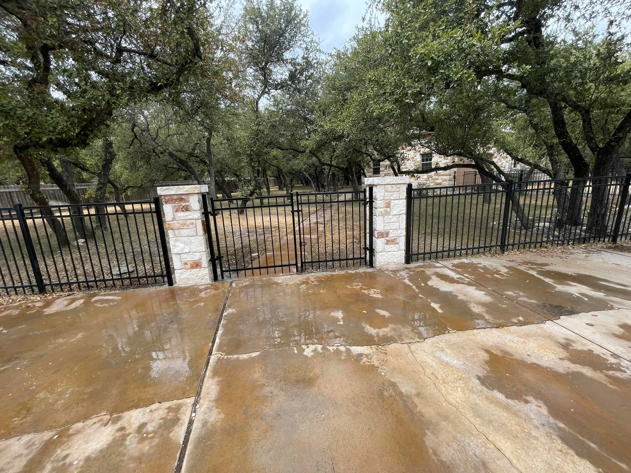 Wet concrete sidewalk in front of a black metal fence with stone pillars, leading to a yard with trees and a house in the background.