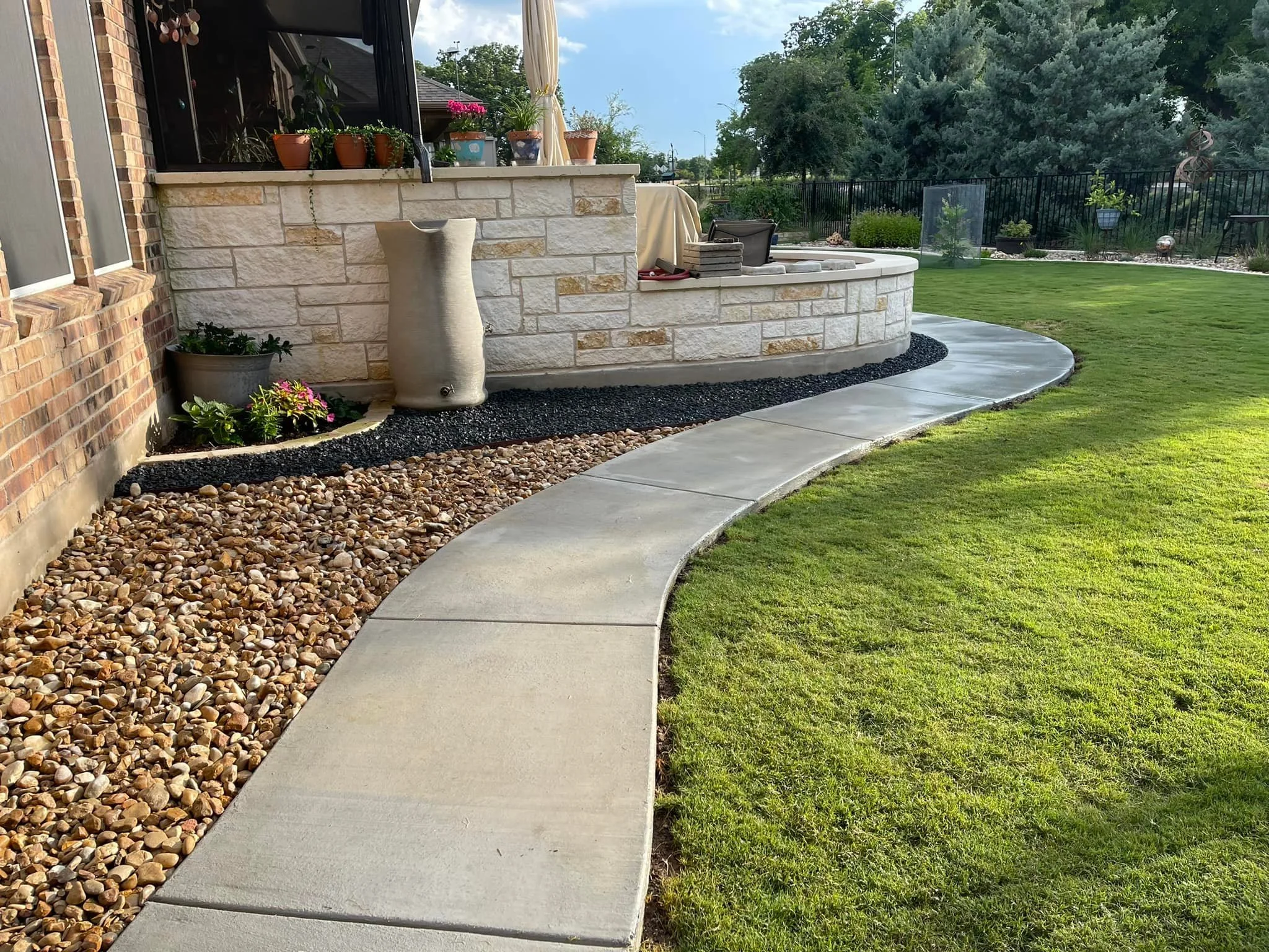A curved concrete garden pathway runs alongside a well-manicured lawn, with decorative rocks and small plants edging the path near a house with a brick and stone exterior. There are potted plants and outdoor decor on the porch area.